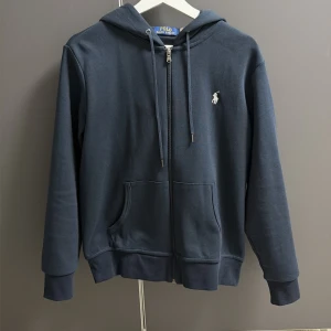 Marinblå hoodie från Polo Ralph Lauren - Snygg marinblå hoodie från Polo Ralph Lauren med dragkedja och huva. Tröjan har snörning vid halsen, två fickor fram och den klassiska broderade loggan på bröstet. Perfekt för chill dagar och enkel att matcha med jeans eller joggers.