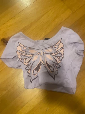 Ljuslila croppad topp med fjäril - Superfin ljuslila croppad topp från Shein med ett stort tryck av en fjäril i rosa och brunt på framsidan. Toppen har korta ärmar och en bred, något bågformad halsringning. Perfekt för dig som gillar statement-plagg och pastellfärger.