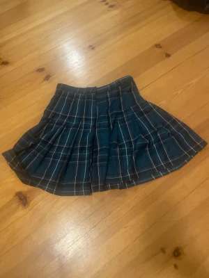 Mörkblå rutig plisserad kortkjol - Säljer en mörkblå plisserad kortkjol med klassiskt rutigt mönster i vitt och grått. Kjolen har en snygg midjeform och ger en cool skoluniform vibe. Perfekt för dig som gillar preppy stil och vill sticka ut med en trendig look.