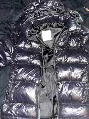 Svart pufferjacka från Moncler - Säljer en svart pufferjacka från Moncler med glansig finish och klassisk quiltad design. Jackan har huva, två sidofickor med dragkedja och coola Moncler-detaljer på dragkedjorna. Insidan har en unik serietidningspatch som ger extra vibe.