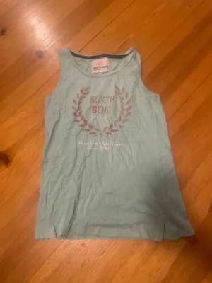 Ljusgrön linnetopp från North Bend - Snygg ljusgrön linnetopp från North Bend med tryckt logga och krans i rött framtill. Toppen är ärmlös och har en klassisk rak passform. Perfekt för varma dagar och enkel att matcha med jeans eller shorts.