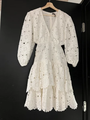 maje dress - Superfin vit kortklänning från Maje med v-ringning, långärmade puffärmar och snygga cut-outs i midjan. Klänningen har ett detaljerat broderat spetsmönster och volanger i kjolen. Dragkedja i ryggen för enkel påklädning. Perfekt för dig som gillar romantisk och trendig stil.