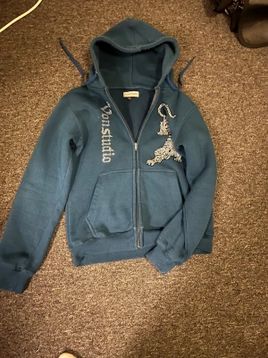 Blå hoodie med tigertryck från Vonstudio - Snygg mörkblå hoodie från Vonstudio med huva och dragkedja framtill. På bröstet finns ett coolt tigertryck i silver och texten 'Vonstudio' i glittriga bokstäver. Tröjan har långa ärmar och två fickor framtill. 
