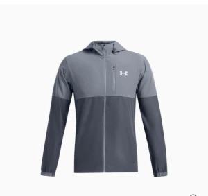Grå vindjacka från Under Armour - Snygg tvåtonad grå vindjacka från Under Armour med huva och hel dragkedja framtill. Jackan har långärmade ärmar med elastiska muddar och en diskret logga på bröstet. Perfekt för dig som gillar sportig stil och funktionella plagg.