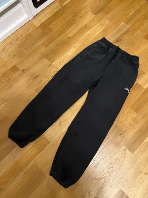 Svarta mjukisbyxor från Stüssy XS - Svarta mjukisbyxor från Stüssy i storlek XS. Byxorna har resår och snörning i midjan, samt Stüssy-logga broderad på benet. Tillverkade i mjuk bomull och polyester, med ribbade muddar vid bensluten. Perfekta för chill och streetwear.