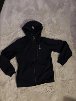 Mörkblå hoodie från Sail Racing - Mörkblå hoodie från Sail Racing med hel dragkedja och snörning i huvan. Jackan har en bröstficka med dragkedja och logotyp på både bröstet och huvan. Mjuk bomullskänsla och långärmad modell, perfekt för chill dagar.