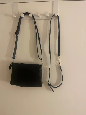 Svart axelväska med gulddetaljer - Stilren svart axelväska i skinn med flera fack och dragkedjor i guld. Väskan har justerbar rem och en strukturerad, lite boxig form. 