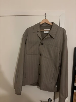Beige overshirt från Les Deux - Snygg beige overshirt från Les Deux med diskret fiskbensmönster. Jackan har klassisk krage, stora bröstfickor och knäppning med svarta knappar. Perfekt att slänga över en hoodie eller t-shirt för en chill look.