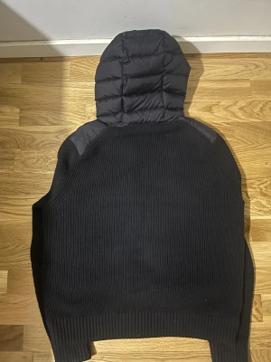 Svart Moncler pufferjacka med huva - Svart cardigan  från Moncler med stickade ärmar och rygg, quiltad framsida och huva. Jackan har tvåvägsdragkedja, ribbade muddar och Moncler-logga på ärmen. Materialmix av polyester och stickad textil ger en unik look. Perfekt för dig som gillar streetstyle. Kvitto finns. 