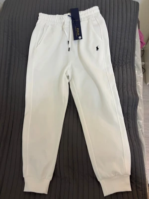 Vita mjukisbyxor från Polo Ralph Lauren - Snygga vita mjukisbyxor från Polo Ralph Lauren med klassisk logga broderad på benet. Byxorna har elastisk midja med snörning, ribbade muddar vid benslut och två sidofickor. Byxorna är L men passar m