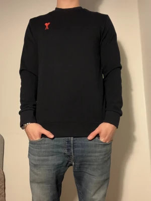 Svart sweatshirt från Ami - Snygg svart sweatshirt från Ami i storlek M. Tröjan har ett broderat rött hjärta med bokstaven A på bröstet, rund hals och långa ärmar. Tillverkad i mjuk bomull med normal passform, perfekt för en chill och stilren look. 