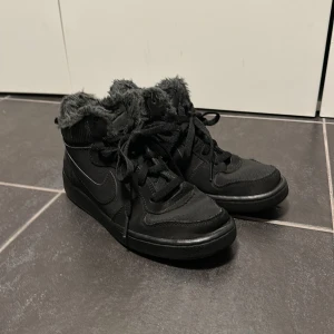 Svarta Nike sneakers med pälsfoder - Svarta Nike sneakers med högt skaft och mjukt grått pälsfoder runt kanten. Skorna har snörning, rund tå och platt sula. Snyggt Nike-märke på hälen och tungan. Perfekta för dig som vill ha både stil och värme under kalla dagar.