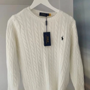 Vit kabelstickad tröja Polo Ralph Lauren - Vit kabelstickad tröja från Polo Ralph Lauren med rund hals och klassisk broderad logga på bröstet. Tröjan har långärmad passform och ribbade muddar vid ärmslut och nederkant. Perfekt för dig som gillar stilrena och tidlösa plagg.
