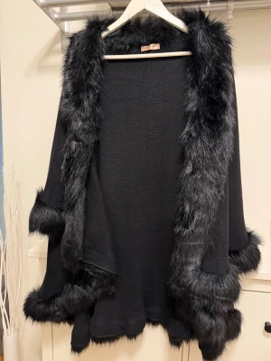 Svart cape med fuskpälsdetaljer - Säljer en svart cape med breda fuskpälsdetaljer längs krage, ärmslut och nederkant. Jackan har en loose passform och ger en lyxig vibe med sin fluffiga pälslook. Perfekt för dig som vill sticka ut med en elegant och mysig stil.