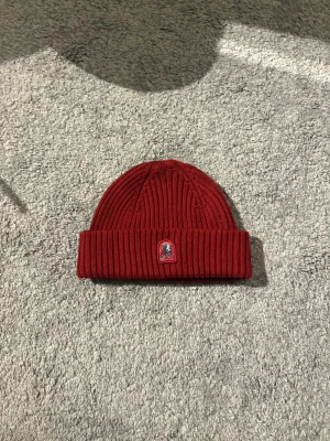Röd parajumper mössa OANVÄND - Snygg röd ribbstickad mössa från Parajumpers med uppvikt kant och klassisk PJS-logga framtill. Tillverkad i mjuk ullblandning som håller dig varm under kalla dagar. Perfekt accessoar för att lyfta din vinterstil.