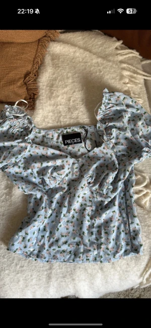 Blommig ljusblå blus från Pieces - Supersöt blus från Pieces i ljusblått med små blommor i rosa och vitt. Modellen har puffiga korta ärmar med volang och knytband vid axlarna. Blusen är figurnära med lätt rynk vid bysten och har en romantisk vibe, perfekt för vår och sommar. Aldrig använd! Dragkedja på sidan