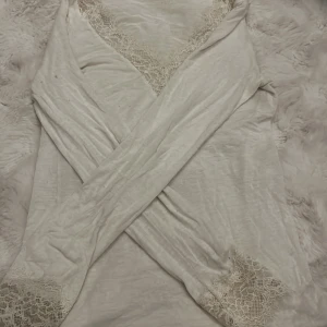 Vit omlottopp intimissimi - Säljer en ljusvit omlottopp med v-ringad hals och fina spetsdetaljer längs ringningen och ärmsluten. Toppen har långärmade armar . Tröjan har liten anmärkning inget som syns så mycket.  Iallfall mjukt, tunt material som känns skönt mot huden. Perfekt för dig som gillar romantiska och feminina vibes.