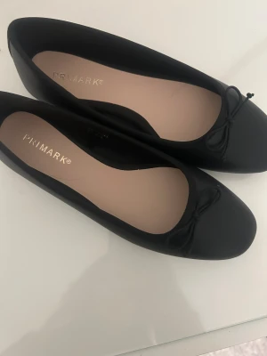 Svarta ballerinaskor från Primark - Stilrena svarta ballerinaskor från Primark med rund tå och en söt rosettdetalj framtill. Skorna har en platt sula och är tillverkade i syntetmaterial, vilket gör dem lätta och bekväma att bära hela dagen.