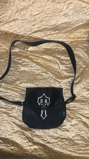 Svart axelväska från Trapstar - Svart axelväska från Trapstar med justerbar rem där loggan är invävd. Väskan har ett coolt vitt emblem på locket och ett spänne framtill. Materialet är slitstark polyester och designen är stilren med mesh-detaljer på framsidan.