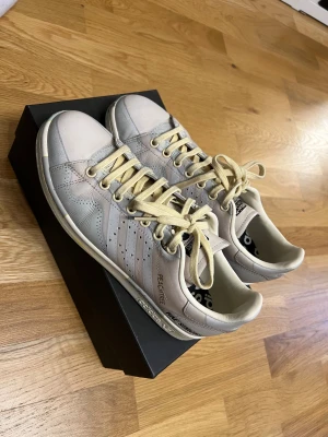 Adidas Stan Smith Raf Simons sneakers - Unika Adidas Stan Smith Raf Simons sneakers i ljusbeige och vitt skinn med krämfärgade snören. Snygga detaljer som perforerade ränder, texten 'PEACHTREE' och 'RAF SIMONS' på sidan samt subtila gula linjer på sulan. Klassisk rund tå och platt sula.