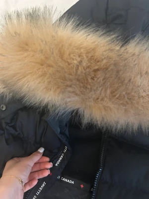 Svart pufferjacka med päls från Zavetti Canada - Svart pufferjacka från Zavetti Canada i storlek XS med fluffig beige päls på huvan. Jackan har flera dragkedjor, detaljer med röda sömmar och ett snyggt märke på ärmen. Perfekt för kalla dagar med sin tjocka och isolerande design.