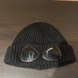 Svart C.P. Company Goggle mössa OBS KAN PRUTAS! - Svart ribbstickad mössa från C.P. Company med ikoniska goggle-linser framtill. Mössan är tillverkad i mjuk ull och har en uppvikt kant. Perfekt för dig som vill sticka ut med en streetwear-vibe och gillar detaljer som syns. KAN PRUTAS! Aldrig använd!