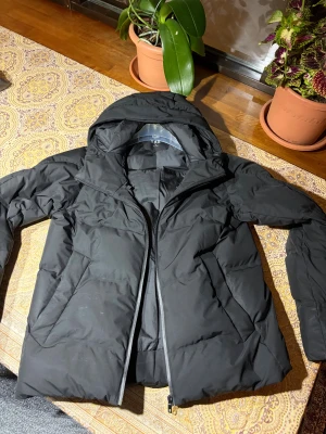 Svart pufferjacka från Uniqlo M - Svart pufferjacka från Uniqlo i storlek M. Jackan har huva, dragkedja och två sidofickor. Materialet är lätt, vindtätt och vattenavvisande syntet. Perfekt för kalla dagar med sin quiltade design och stilrena look.