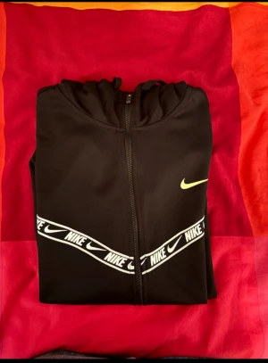 Svart Nike vindjacka med huva M - Nike vindjacka i svart med huva och hel dragkedja. Snygga vita Nike-loggor på bröstet och diagonalt band över framsidan. Jackan har lång ärm och är gjord i polyester, perfekt för sportiga dagar. Ikonisk gul Nike Swoosh på bröstet för extra stil.