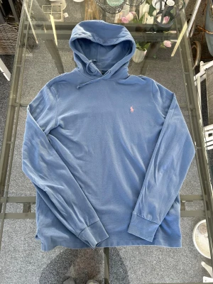 Polo Ralph Lauren blå hoodie M – klassisk logga & fint skick - Snygg Polo Ralph Lauren hoodie i en dämpad ljusblå färg, storlek M. Mjuk och tunnare bomullskvalitet som gör den perfekt för vardag, vår/sommar eller som ett lager under jacka. Ikonisk rosa Polo-brodyr på bröstet. Äkta vara – se etikettbild.  🧵 Passform: Regular fit 🎨 Färg: Ljusblå / washed blue 📏 Storlek: M ✨ Skick: Mycket bra, inga fläckar eller skador  Ett klassiskt och tidlöst plagg för dig som gillar premiumkänsla och ren stil. 