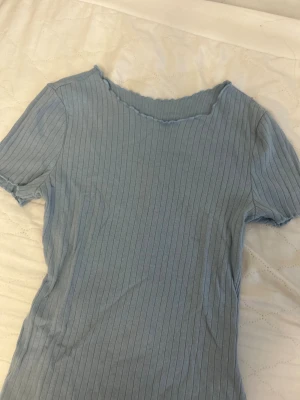 Ljusblå ribbad topp med volangkant - Säljer en ljusblå ribbad topp med korta ärmar och volangkant vid halsringning, ärmslut och nederkant. Toppen har en slim passform och är tillverkad i mjuk bomull. Perfekt för dig som gillar enkel men snygg stil.