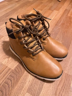 Bruna boots med snörning från Everest - Snygga bruna boots från Everest med svart detalj vid hälen och vit sula med svarta prickar. Skorna har rund tå, snörning och är tillverkade i skinn och textil. Perfekta för höst och vinter, med en cool streetkänsla och robust look.