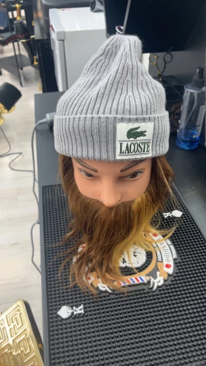 Grå ribbad mössa från Lacoste - Säljer en snygg grå ribbstickad mössa från Lacoste med klassisk logotyp framtill. Mössan är unisex och tillverkad i 100% ull, vilket gör den både mjuk och varm. Toppen för dig som vill vara riktigt grisch.