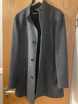 Grå rock från Riley, storlek 52 - Snygg grå rock från Riley med klassisk rak passform och ståkrage. Jackan har knäppning med svarta knappar och två sidofickor. Insidan har ett quiltat foder med dragkedja för extra värme, även avtagbar. Ursprungs pris 3000kr. Inga defekter finns på jackan.