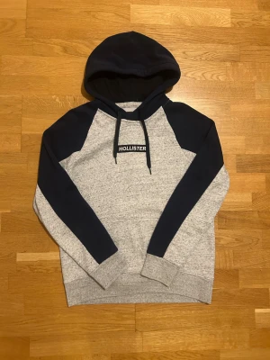Grå och marinblå hoodie från Hollister - Snygg hoodie från Hollister i storlek S med gråmelerad kropp och marinblå ärmar och huva. Tröjan har snörning vid huvan och ett broderat Hollister-tryck på bröstet. Perfekt för chill dagar och streetstyle. Mjuk insida och ribbade muddar för extra komfort.