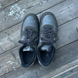 Nike Air Force 1 i grå mocka - Säljer ett par Nike Air Force 1 i grå mocka med svarta detaljer och svart sula. Skorna har klassisk siluett, snörning och rund tå. Swoosh-loggan i ljusgrått på sidorna och Nike Air-brodyr på hälen. Perfekta för dig som gillar streetstyle och tidlös design.