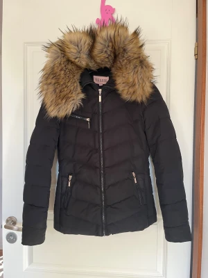 Svart jacka med fuskpäls luva från Tessie - Svart jacka från Tessie i storlek 32. Jackan har en stor fluffig (supermjuk) fuskpäls som ser äkta ut i bruna toner längst med luvan. Den har tre dragkedjeförsedda fickor fram och markerad midja som formar kroppen snyggt. Perfekt vinterjacka!