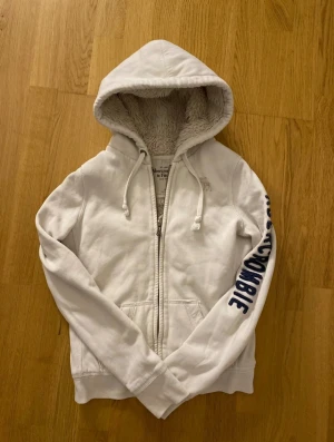 Vit zip hoodie Abercrombie & Fitch - Mysig vit hoodie från Abercrombie & Fitch med fluffigt foder på insidan och huva med snörning. Tröjan har dragkedja framtill och tryckt text på ena ärmen. Storlek M men passar som S. Längd 55cm, Bredd 45cm