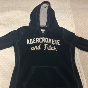 Mörkblå hoodie Abercrombie & Fitch  - Snygg mörkblå hoodie från Abercrombie & Fitch med stor vit logotyp framtill. Tröjan har huva med snörning, känguruficka och långa ärmar. Perfekt för chill dagar och enkel att matcha med jeans eller leggings. Storlek M men passar bättre till S&XS. 