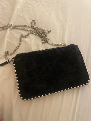 Svart mocka axelväska med kedja från Zara - Svart axelväska i mocka med silverfärgade kuldetaljer runt kanterna och en lång kedja som axelrem. Väskan har flera fack och dragkedja upptill.