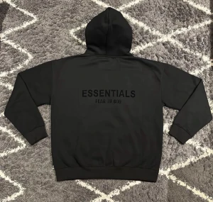 Svart Essentials Fear of God hoodie - Svart hoodie från Essentials Fear of God med stor logga på ryggen och mindre logga framtill. Tröjan har huva, känguruficka och ribbade muddar. Tillverkad i mjuk bomull och polyesterblandning, perfekt för en chill och stilren look.