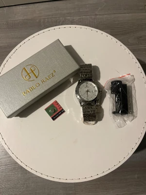 Silverfärgad klocka från Pablo Raez:  - Stilren klocka från Pablo Raez med rund urtavla i silver och romerska siffror. Armbandet är i blankt metall och har en länkdesign. Klockan har datumvisning och levereras med originalask, batteri och verktyg. Perfekt accessoar för dig som gillar klassisk look.