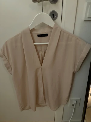 Beige blus med v-ringning från Vila - Säljer en stilren beige blus från Vila i storlek S. Blusen har v-ringad hals, korta ärmar och ett avslappnat fall. 