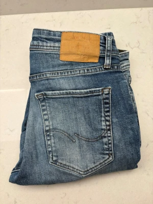 Blå jeans från Jack & Jones - Snygga blå jeans från Jack & Jones med klassisk femficksdesign och läderpatch bak i midjan. Jeansen har en tvättad look med lätt slitning och raka ben. Perfekt för dig som gillar en avslappnad och tidlös stil.