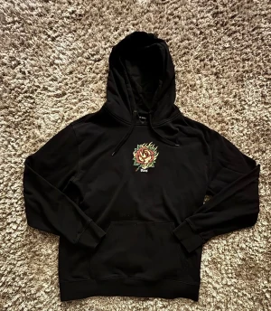 Svart Vans hoodie med ros-tryck - Svart hoodie från Vans i storlek L med en fet ros och flames-tryck på bröstet. Klassisk känguruficka och dragsko i huvan. Mjuk insida och cool streetstil. Jag har inte använt den något då den är för stor för mig.Perfekt för dig som gillar unika prints och skatevibe. Har du några frågor så är det bara att skriva.