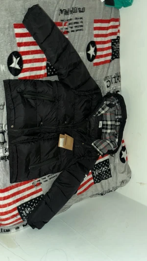 Svart Burberry pufferjacka med huva - Svart pufferjacka från Burberry med avtagbara ärmar, vilket gör att den kan användas som väst. Jackan har en stor huva med klassiskt rutigt Burberry-foder i svart, grått och vitt. Två dragkedjefickor framtill och tryckknappar. Snyggt Burberry-tryck på ena ärmen.