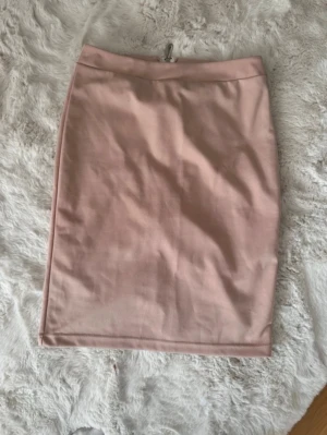 Ljusrosa pennkjol med dragkedja - Säljer en stilren ljusrosa pennkjol med smal passform och dragkedja bak. Kjolen är kort och har en enkel, slät design utan mönster eller extra detaljer. Perfekt för dig som gillar minimalistisk och feminin stil.