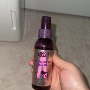 Aussie Reconstructor Hair Oil - Aussie Reconstructor Hair Oil i en lila plastflaska med pump. Oljan är lättviktig och passar för att ge håret extra glans och näring. Flaskan har snyggt tryck med rosa och lila detaljer samt en känguru på etiketten. Perfekt för dig som vill boosta håret med fukt och mjukhet.