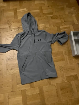 Grå vindjacka från Under Armour - Grå vindjacka från Under Armour med huva och svart logga på bröstet. Jackan har hel dragkedja framtill, två sidofickor och elastiska muddar vid ärmsluten. Perfekt för träning och aktiva dagar utomhus.