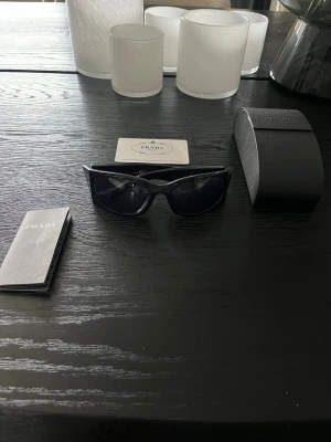 Svarta solglasögon från Prada - Snygga svarta solglasögon från Prada med breda skalmar och mörka linser. Glasögonen har en stilren, rektangulär form och kommer med originalfodral, certifikat och manual. Tillverkade i Italien och har Prada-logga på skalmen.