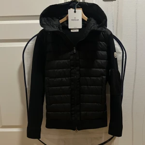 Moncler Cardigan - OBS: nfc funkar ej pga nyare modell. Svart pufferjacka från Moncler med huva och dragkedja framtill. Jackan har quiltad front i syntetmaterial och stickade ärmar med Moncler-logga på vänster arm. Praktiska fickor med knappstängning och ribbade muddar. Perfekt för kyliga dagar.